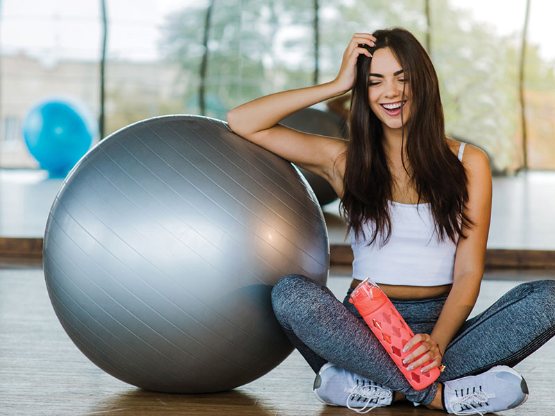 lady sitting on the gym floor smiling with a water bottle leaning on a medicine ball